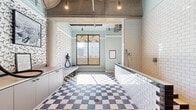 Located in Baltimore, MD, this thoughtfully designed pet spa delivers a distinctive blend of practicality and playful character. The single-floor space is efficiently organized, making the most of its compact footprint through purposeful layout and eye-catching design choices.

The flooring throughout is a standout feature — bold checkered tiles in blue-and-white and black-and-white patterns lend a retro yet polished aesthetic. The walls are adorned with charming dog-print wallpaper that wraps the space in pet-friendly personality, complemented by soft mint-green painted surfaces that create a clean, welcoming ambiance.

A tiled grooming and wash station anchors one side of the space, constructed with classic white subway tiles and accessible step-up entry. Built-in cabinetry with brass hardware provides organized storage for grooming tools and supplies. A wall-mounted dryer and leash hooks add functional touches to the grooming area.

Natural light enters through a large window overlooking the Baltimore cityscape, with a "Happy Pets, Happy Life" graphic decal adding a branded, cheerful note. The exposed concrete ceiling with industrial pendant lighting gives the space an urban loft quality. Framed pet photography and informational postings complete the interior, balancing function with a warm, inviting atmosphere for pets and their owners alike.