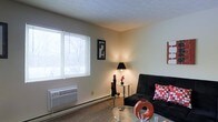 This single-floor unit offers a practical and comfortable living environment with a welcoming ambiance. The living area is finished with wall-to-wall carpeting that adds warmth underfoot and is illuminated by a large window fitted with horizontal blinds, allowing an abundance of natural light to fill the room while maintaining privacy. A wall-mounted air conditioning and heating unit ensures year-round climate control with ease and convenience. The living space is designed to accommodate a comfortable seating arrangement, as evidenced by the layout's suitability for a sofa and glass-top coffee table combination. A baseboard heater runs along the exterior wall, providing supplemental warmth during cooler months. The neutral-toned walls provide a versatile backdrop, allowing for a variety of décor styles. A floor lamp adds a soft, ambient glow to complement the natural light. Overall, the space presents a straightforward and functional layout well-suited for everyday living, with key features that prioritize comfort and practicality throughout.