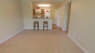 This single-floor space offers an open-concept layout that integrates the living area and kitchen into one cohesive, functional environment. The living area features plush wall-to-wall carpeting in a warm neutral tone, creating a comfortable and inviting atmosphere. Clean white baseboards and softly painted walls contribute to the bright, airy feel of the space.

The kitchen is positioned at the back of the open floor plan and is accessible via a raised breakfast bar counter, which accommodates two woven bar stools — a practical and stylish touch for casual dining or entertaining. The kitchen is equipped with white cabinetry featuring open upper shelving, providing convenient storage and display space. A full-size white refrigerator anchors one side of the kitchen, and the countertops offer ample prep space.

A hardwood or wood-look flooring transitions from the kitchen area toward a hallway, which leads to additional rooms and provides a natural delineation between the living and utility spaces. A doorway visible from the main living area hints at further rooms beyond. Overhead fluorescent lighting in the kitchen ensures the space is well-lit, while the overall design emphasizes a clean, functional aesthetic throughout.