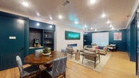 This thoughtfully designed single-floor space blends vibrant personality with comfortable functionality, making it an inviting destination for socializing and relaxation. The interior is anchored by a striking deep teal color palette applied to the walls, built-in shelving, and wainscoting details, creating a bold yet cohesive aesthetic throughout.

The lounge area serves as the centerpiece of the space, featuring a wall-mounted neon "Stay Awhile" sign set against a lush moss panel backdrop, positioned above a sleek horizontal electric fireplace. This focal wall is flanked by symmetrical dark teal built-in bookshelves, adding both storage and visual depth. Comfortable seating — including a floral-patterned sofa, upholstered lounge chairs, and a wood-and-metal coffee table — is arranged atop a patterned area rug, encouraging casual conversation and relaxation.

Adjacent to the lounge is a defined dining area, centered around a warm-toned wooden round table surrounded by fabric chairs upholstered in a botanical print. A self-serve beverage station with a coffee machine is conveniently integrated into the nearby built-in cabinetry. Framed artwork decorates the teal walls, adding a curated, gallery-like quality to the space.

Light hardwood flooring runs throughout, complemented by recessed lighting and a classic beadboard ceiling that adds texture and a touch of coastal charm to the overall design.