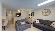 This single-floor residence showcases a thoughtfully designed open-concept layout that seamlessly blends the living area and kitchen into one cohesive space. The living room is warm and inviting, featuring plush carpeted flooring and a neutral color palette that creates a calm, welcoming ambiance. Two generously sized gray sofas anchor the room, complemented by tasteful decorative accents including wall clocks and a classic whitewashed cabinet that adds a charming focal point. A skylight panel in the ceiling allows natural light to fill the space, enhancing its airy feel alongside the flush-mount and pendant lighting fixtures overhead. The open-plan kitchen is visible beyond a half-wall breakfast bar, outfitted with dark cabinetry that provides a striking contrast to the light walls and countertops. This pass-through design between the kitchen and living area promotes connectivity and ease of entertaining. The overall aesthetic is clean and contemporary, with soft tones and thoughtful touches throughout that make the space feel both functional and comfortable for everyday living.