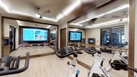 This dedicated fitness studio is thoughtfully designed to deliver a professional, immersive workout experience. The space centers around a row of Spinning stationary bikes arranged on a light hardwood floor, offering an energetic cycling environment. Two large flat-screen display monitors — framed in warm wood trim and mounted prominently — provide interactive workout programming and visual motivation during sessions. Wall-to-wall mirrors expand the sense of space and allow participants to monitor their form, while also reflecting the room's clean, modern aesthetic throughout. A classical ballet barre runs along one mirrored wall, adding versatility to the studio for barre classes and stretching exercises beyond cycling. The ceiling features recessed linear lighting paired with ceiling fans, ensuring the space remains well-lit and comfortable during even the most intense workouts. Neutral tones in the walls and flooring are complemented by rich wood accents, lending the studio a warm yet contemporary feel. The overall ambiance is polished and purpose-built, blending practical fitness functionality with an inviting, upscale atmosphere that encourages consistent use and a premium exercise experience.