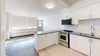 Located at 4750 Bathurst Street in Toronto's North York neighbourhood, this ground-floor unit offers a practical and efficiently designed single-floor layout. The open-concept kitchen is a standout feature, outfitted with crisp white cabinetry, a classic white subway tile backsplash, dark countertops, and a full suite of stainless steel appliances including a range, over-the-range microwave, and refrigerator. A peninsula counter separates the kitchen from the adjoining living area, creating a natural flow between the two spaces. The living area is bright and airy, bathed in natural light through a large window fitted with a roller blind, and features classic parquet wood flooring that adds warmth and character to the clean white walls. A baseboard radiator provides in-unit heating. Flush-mount ceiling lighting illuminates each area consistently. The unit includes a private entry door visible from the interior. Outside, the property offers access to a paved parking area framed by mature trees and green landscaping, with open street views along Bathurst Street. The overall ambiance is clean, functional, and well-maintained, making it a practical urban unit in a well-established Toronto neighbourhood.