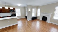 This single-floor residence blends classic architectural charm with modern functionality. The open-concept living area is anchored by rich hardwood flooring that extends throughout, complemented by crisp white crown molding and clean baseboards that add a timeless elegance to the space. Warm neutral-toned walls create a welcoming ambiance that pairs well with the abundant natural light streaming through multiple white-framed windows fitted with horizontal blinds.

A standout feature of the living area is the classic decorative fireplace with a white surround and dark hearth, serving as a strong focal point. A traditional cast-iron radiator beneath one of the windows adds to the space's vintage character. A wall sconce provides additional ambient lighting, enhancing the room's warmth.

The kitchen is seamlessly integrated into the open layout, offering stainless steel appliances including a range and an over-the-range microwave, as well as a light-colored countertop that provides a functional workspace. The transition between the kitchen and living area is smooth and unobstructed, making the overall space feel expansive and ideal for both everyday living and entertaining.