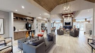 The Lodges On English Station clubhouse is a thoughtfully designed single-story communal space that blends rustic charm with modern sophistication. The layout is open and expansive, with distinct zones flowing seamlessly into one another across light-toned wood-look tile flooring throughout.

The entry lounge area features soaring vaulted ceilings clad in warm wood paneling, illuminated by an elegant wrought-iron chandelier. Tall glass French doors flood the space with natural light and offer views of the surrounding outdoor areas. This section is furnished with plush gray sofas and organic accent pieces, creating a welcoming, relaxed atmosphere.

The central gathering area is anchored by a dramatic fireplace wall, where a linear gas fireplace is framed by dark metal and reclaimed wood detailing, with a large wall-mounted TV above. Exposed dark wood ceiling beams and industrial-style pendant lighting define this zone, complemented by multiple seating arrangements in neutral and rust tones.

Adjacent to the lounge is a well-appointed bar and kitchenette area featuring a stone tile backsplash, an extended counter with bar stools, pendant lighting, a beverage refrigerator, and ample cabinetry. A game room with a billiards table is also visible, adding a recreational dimension. Together, these areas create a versatile, inviting communal environment.