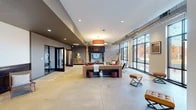 This single-floor amenity space combines industrial and contemporary design across several distinct areas. The expansive main lounge serves as the centerpiece, featuring polished concrete floors, exposed metallic ductwork running along the ceiling, and floor-to-ceiling steel-framed windows that flood the space with natural light and offer views of the surrounding outdoors. A brick accent wall frames a built-in fireplace with a mounted television above, while a bold orange drum pendant light anchors the seating area furnished with rustic wooden tables and leather ottomans.

Glass-partitioned offices and a conference room branch off from the lounge, the latter highlighted by a rich walnut conference table, modern task chairs, and a distinctive vintage "Colosseum Entertainment Center" banner adorning the wall. An entry vestibule with large glazed doors and blue carpeting provides a clean, functional transition point into the building.

The well-equipped fitness center is bathed in natural light from expansive windows overlooking tree-lined grounds. It features multiple treadmills, an elliptical, weight benches, free weights, and full-length mirrors, with exposed ductwork overhead reinforcing the industrial aesthetic. A vibrant branded mural reading "Home Is Here" adds an energetic focal point to the cardio area.