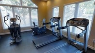 This dedicated fitness space is designed to support an active lifestyle, offering a focused environment for cardio training and exercise. The room is outfitted with multiple pieces of cardio equipment, including treadmills and a recumbent stationary bike, providing a variety of workout options for users of different fitness levels. Durable rubber flooring covers the entire area, offering both safety and comfort during exercise sessions. Large windows line the walls, allowing generous amounts of natural light to fill the space and offering pleasant views of surrounding greenery and trees — creating a refreshing, open atmosphere that enhances the workout experience. White horizontal blinds on each window provide the option for light control and privacy as needed. The warm, golden-toned walls contribute to an energizing yet welcoming ambiance. The layout is thoughtfully arranged to allow ample space around each piece of equipment, ensuring ease of movement and safe use. The room is single-level, straightforward in its design, and purpose-built for fitness, making it a practical and functional amenity for regular use.