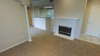 This single-floor space offers an open-concept layout that connects the living area and kitchen in a seamless flow. The living room is anchored by a white-mantled fireplace, providing a classic focal point, and features a decorative column that adds a touch of architectural character. Neutral-toned carpeting spans the living area, contributing to a warm and understated ambiance throughout.

The kitchen is positioned adjacent to the living space and is partially separated by a raised breakfast bar with a dark countertop, offering both a casual dining option and a natural visual boundary between the two areas. White cabinetry lines the kitchen walls, complemented by white appliances including a microwave and range, lending the space a clean, cohesive appearance. Overhead fluorescent lighting illuminates the kitchen workspace effectively.

A hallway visible beyond the living area suggests additional rooms further into the unit. The overall color palette — consisting of beige walls, white trim, and neutral flooring — creates a consistent, move-in-ready aesthetic throughout the space.