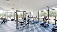 Located at 254 Lynnway in Lynn, Massachusetts, this contemporary fitness center is thoughtfully designed to support a full range of workout routines. The facility is organized into two distinct areas: a main gym floor and a dedicated studio space.

The main gym floor features a striking geometric-patterned carpet in shades of blue, white, and teal, lending the space a vibrant, modern aesthetic. Floor-to-ceiling windows line the perimeter, flooding the room with natural light and offering unobstructed views of the surrounding greenery. Overhead, large circular pendant lights provide additional illumination and contribute to the space's clean, contemporary design. Equipment includes a full rack of dumbbells, a weight bench, a multi-function cable and power rack system, multiple TRUE-brand treadmills, elliptical cross-trainers, and a stationary air bike.

The studio area features warm hardwood flooring and a full wall of mirrors, creating an open, bright environment ideal for group classes, stretching, yoga, or personal training sessions. A functional training station with TRX suspension equipment, medicine balls, and kettlebells is positioned along one wall, offering versatile training options. Built-in shelving provides organized storage for accessories. The studio connects directly to the main gym floor through glass-paneled doors, maintaining a sense of openness throughout the facility.