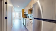 This single-floor unit showcases a thoughtfully designed layout with modern finishes throughout. The kitchen is a standout feature, equipped with stainless steel appliances including a full-size refrigerator, dishwasher, over-the-range microwave, and electric range. Light wood cabinetry with sleek black hardware lines both the upper and lower sections, complemented by clean white countertops. A deep single-basin undermount sink with a matte black pull-down faucet anchors the workspace, while a striking hexagonal teal tile backsplash adds a distinctive design element. Recessed lighting illuminates the space efficiently.

The bathroom continues the modern aesthetic with a wood-toned vanity featuring drawer and cabinet storage, white countertop, a large frameless mirror, and matte black hardware accents. The flooring carries a subtle stone-like appearance, adding texture to the clean, neutral palette.

The living and sleeping areas benefit from large floor-to-ceiling windows that flood the space with natural light and provide open views of the surrounding urban landscape. Light wood-toned flooring runs throughout these areas, contributing to the bright and airy atmosphere. A sliding barn-style door is visible along the corridor, adding a functional yet stylish architectural detail to the overall design.