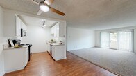 This single-floor unit offers a clean, functional layout designed for comfortable living. The open-concept living and dining area is anchored by warm hardwood-style flooring, a ceiling fan with a light fixture, and a textured ceiling. Large sliding glass doors with vertical blinds lead to a private balcony, filling the space with natural light and providing outdoor views. The galley-style kitchen is well-equipped with white cabinetry, light stone countertops, a black electric range, a built-in microwave, a dishwasher, and a stainless steel sink. Track lighting illuminates the workspace efficiently. Transitioning into the living area, plush neutral carpet extends throughout the carpeted zones, creating a cohesive and comfortable feel. The bedrooms are generously proportioned, each featuring carpet flooring, white walls, a textured ceiling, and windows with vertical blinds that admit ample natural light and views of surrounding greenery. The bathroom is finished with hardwood-style flooring, a vanity with a light stone countertop, a large mirror, and a glass-enclosed shower. Clean white walls and a neutral palette throughout the unit contribute to a bright, airy ambiance that is both practical and inviting.