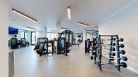This professionally designed fitness center offers a spacious, single-floor layout that accommodates a wide variety of workout disciplines. The space is defined by its clean, contemporary aesthetic — crisp white walls, structural columns, and light wood-tone flooring create a bright, open environment that feels both functional and inviting.

Overhead, linear pendant lighting runs throughout the facility, providing consistent, well-distributed illumination across all workout zones. Large windows along the perimeter allow natural light to supplement the interior lighting, further enhancing the airy atmosphere.

The strength training area is extensively equipped with premium machines from brands including Cybex and Life Fitness, covering chest press, leg press, cable systems, and multi-functional cable stations. A well-stocked dumbbell rack provides a full range of free weights alongside curl bars, catering to both beginner and advanced lifters.

The cardio zone features a row of modern treadmills with individual display screens, as well as stair climbers and stationary cycling bikes, all positioned for ease of use and spacing. A dedicated studio-style area with floor-to-ceiling mirrors, spin bikes, and a foam roller station supports group or individual training sessions.

Flat-screen televisions are mounted throughout the space, and a large wall clock adds practical convenience. The overall design balances high-end equipment density with generous open floor space, making this fitness center a comprehensive and well-appointed training facility.