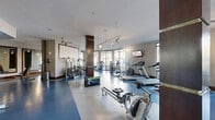 This fitness center occupies a single, open floor plan of substantial size, thoughtfully designed to accommodate a wide range of workout activities. The layout is divided into functional zones, with cardio equipment—including treadmills, ellipticals, and rowing machines—positioned along the perimeter near floor-to-ceiling windows that flood the space with natural light and provide views of the surrounding outdoor greenery.

The strength training area features a variety of cable machines, weight stations, and a multi-functional rack system, complemented by large wall-mounted mirrors that visually expand the space and allow users to monitor their form. Dark wood-clad structural columns add an architectural contrast against the neutral gray walls and textured ceiling, lending a polished, contemporary aesthetic throughout.

Flooring consists of durable, two-tone blue and gray fitness flooring, providing both functionality and visual definition between workout zones. Overhead lighting combines ceiling fans, pendant lights, and adjustable track spotlights, ensuring well-distributed illumination across all areas. A wall-mounted television and a dedicated storage cubby unit near the entrance enhance convenience. The space also incorporates a mirrored accent wall that amplifies depth and reinforces the gym's open, airy ambiance.