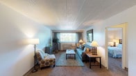 This single-floor residence is thoughtfully laid out to maximize functionality and comfort across its key living areas. The living room serves as the central gathering space, featuring wall-to-wall carpeting, a textured tile ceiling, and a large window dressed with draping curtains that invite generous natural light and outdoor views. The room offers ample space for seating arrangements and connects openly to a dining area, which accommodates a bistro-style table and chairs. Adjacent to the dining space, the kitchen is efficiently designed with warm wood cabinetry, tile flooring, a double-basin sink, electric range, built-in microwave, and a full-size refrigerator — providing practical storage and workspace. A pass-through counter connects the kitchen to the main living area, enhancing the open feel. The bedroom is a well-proportioned, carpeted retreat featuring a double closet with sliding doors and a window that provides natural light and outdoor views. The bathroom is clean and functional, complete with a single-sink vanity with white cabinetry, a mirrored medicine cabinet, tiled flooring, and a shower-tub combination. Throughout the residence, neutral tones and consistent finishes create a cohesive, understated ambiance.