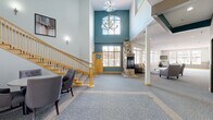 Located at 15734 Foliage Ave in Apple Valley, Minnesota, this community clubhouse offers a welcoming and thoughtfully designed gathering space across a single, generously sized floor. The grand entrance lobby immediately impresses with soaring two-story ceilings, a teal accent wall, and an elegant chandelier that draws the eye upward. A striking floor-to-ceiling stone fireplace anchors the main lounge area, which is furnished with comfortable seating arranged to encourage conversation and relaxation. Soft, patterned carpeting and warm yellow trim details add a cohesive and inviting aesthetic throughout. Large windows flood the space with natural light and provide views of the surrounding outdoor patio area, which is accessible through glass double doors. A staircase with warm wood banisters leads to the upper level, while a passenger elevator ensures convenient accessibility for all residents. The fully equipped kitchen features light maple cabinetry, dark granite countertops, mosaic tile backsplash, and a full suite of stainless steel appliances including a built-in double oven, refrigerator, and dishwasher. A central island with bar seating makes the kitchen ideal for catered events or community gatherings. The overall ambiance is bright, modern, and functional — a refined space designed for community connection.