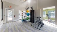 This single-story fitness center offers a thoughtfully designed workout environment with a clean, modern aesthetic. The space is characterized by soaring vaulted ceilings that create an open, airy atmosphere, complemented by neutral gray walls and patterned carpet flooring in grey and chartreuse tones throughout.

Large windows line the perimeter, flooding the interior with abundant natural light and providing views of the surrounding outdoor pool area and landscaping. The layout is organized into distinct functional zones separated by structural columns and open archways. The cardio area houses elliptical trainers and treadmills positioned to take advantage of the poolside views, while the strength training section features a multi-function cable machine, a tiered dumbbell rack with a full range of free weights, and a flat workout bench. A well-stocked medicine ball rack with multiple sizes and colors rounds out the functional training options.

A separate enclosed room, visible through interior glass windows and a glass-panel door, appears to serve as a private studio or meeting space. A ceiling fan overhead and ample ventilation ensure comfort throughout workouts. The overall ambiance is bright, clean, and purposefully arranged to accommodate a variety of fitness routines.