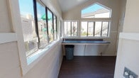 This shared laundry facility is thoughtfully designed to accommodate the needs of a residential community. The room features a striking vaulted ceiling with clerestory windows that allow abundant natural light to fill the space, creating a bright and airy atmosphere. Dark hardwood-style flooring runs throughout, offering a clean and durable surface underfoot.

Multiple front-loading and top-loading washing machines and dryers are arranged along one wall, providing ample capacity for community use. A long, dark-surfaced folding counter stretches beneath a generous bank of windows that frame views of the surrounding community, including a playground area and neighboring buildings. Open shelving above the machines offers convenient storage for laundry baskets and supplies.

The walls feature a classic wainscoting detail in white, complementing the neutral warm-toned upper walls for a tidy, well-maintained appearance. A separate door leads to additional storage or utility space, while posted signage keeps residents informed of usage rules and guidelines. The layout is open and efficient, allowing easy movement between machines and the folding area, making this laundry room a practical and pleasant shared amenity.