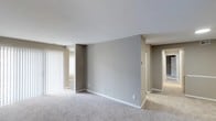 This single-floor space presents a straightforward, functional layout with a clean and neutral aesthetic throughout. The main living area is finished with wall-to-wall carpeting in a light grey-beige tone, contributing to a soft, comfortable underfoot feel that extends into the adjoining spaces visible from the main room.

The walls are painted in a muted grey, complemented by crisp white trim and baseboards that provide subtle contrast. A central architectural dividing wall separates the main living area from a hallway corridor, which leads further into the unit, suggesting additional rooms beyond. This layout creates a natural sense of separation between living and private spaces while maintaining an open sightline.

Natural light enters the space through a large window fitted with vertical blinds on the left side of the main room, brightening the interior and enhancing the airy feel of the open layout. The smooth white ceiling adds to the sense of openness.

The hallway to the right leads to what appears to be additional rooms, with doorways visible in the distance. Overall, the space offers a clean, neutral canvas well-suited for a variety of furnishing styles and uses.