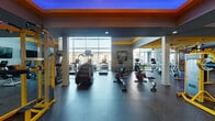 This expansive fitness center in North Little Rock, Arkansas, is designed to deliver a comprehensive workout experience within a stylish, contemporary setting. The facility's bold design palette — featuring striking yellow and gray accents, tray ceilings bathed in ambient blue lighting, and decorative ceiling details — creates an energetic and motivating atmosphere throughout the space.

The cardio zone is a standout feature, lined with Life Fitness treadmills, stationary bikes, stair climbers, and rowing machines. A dramatic floor-to-ceiling glass window wall floods this area with natural light while offering sweeping views of the outdoor pool and surrounding community, making workouts visually engaging.

The strength training area is generously outfitted with a full complement of Life Fitness cable machines, weight racks, free weights, and multi-station resistance equipment, all finished in the facility's signature yellow, providing ample space for varied workout routines. Wall-mounted flat-screen TVs are positioned throughout for entertainment.

Rubber flooring spans the entire facility, providing durability and safety. Ceiling fans, recessed lighting, and chandelier-style fixtures contribute to a well-lit and comfortable environment. Decorative artwork and sports-themed wall pieces add personality to the space. The overall design balances function and aesthetics, making this fitness center a top-tier amenity.