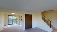 This two-story space offers a functional and straightforward layout with a calm, neutral aesthetic. The main living area is finished with wall-to-wall carpet in a neutral tone, contributing to a quiet and comfortable atmosphere. Light yellow walls run throughout, lending a soft, understated warmth to the interior.

A prominent dark wood double-door closet serves as a central feature of the main level, providing generous built-in storage. To the left, large sliding glass doors frame a view of mature greenery outside, allowing natural light to filter into the space and creating a pleasant indoor-outdoor connection. A hallway extends from this area, leading to additional rooms beyond.

On the right side of the main level, a staircase with elegant wrought iron railings ascends to the second floor, adding a subtle architectural detail to the otherwise simple interior. The upper level is accessible via this staircase, expanding the overall usable space across two floors.

The space maintains a clean, move-in-ready appearance with smooth ceilings and minimal ornamentation, making it highly adaptable to a variety of interior design preferences.