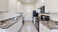 This single-story unit at Jayhawks in Rapid City, South Dakota offers a clean, modern aesthetic throughout, defined by neutral cream-toned walls and light gray luxury vinyl plank flooring that runs consistently across all rooms, creating a cohesive and contemporary feel.

The living area is bright and open, featuring large windows that allow natural light to fill the space while providing views of the surrounding neighborhood. A wall-mounted HVAC unit ensures year-round comfort, and the entry door is positioned conveniently alongside the main window, making for a welcoming and functional entryway.

The galley-style kitchen is well-appointed with white cabinetry, dark granite-style countertops, and a full suite of black appliances including an electric range, microwave, refrigerator, and dishwasher. A rear door with a window provides additional natural light and secondary access.

The bedroom offers generous space and is notable for its dual closets — both featuring bifold doors and built-in hanging rods — providing ample storage. A window overlooks mature trees, adding a natural element to the room.

The bathroom includes a tub-shower combination, toilet, and a white vanity with dark countertop, finished with the same vinyl flooring found throughout the unit. A water heater is also accessible within the bathroom utility area.
