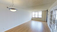 This single-floor space offers a clean, open layout defined by light hardwood flooring that extends throughout the main living area. A large picture window along the far wall serves as a focal point, drawing in generous natural light while framing an elevated urban view of the surrounding cityscape. The window's grid-pane design adds a structured, classic aesthetic to the room. White walls provide a neutral backdrop throughout, complementing the warm tones of the hardwood floors. A pendant light fixture hangs from the textured ceiling, adding a functional yet understated decorative element to the dining or living area. The textured ceiling adds subtle visual character without detracting from the otherwise clean, minimalist interior. Smooth white baseboards trim the perimeter of the room, offering a polished finish. The open floor plan allows for flexible furniture arrangement, making the space adaptable for various living configurations. The elevated position of the unit, suggested by the window views, contributes to a bright and airy atmosphere throughout the day, enhancing the overall sense of openness within the space.