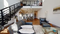 Located at 200 W Grand Ave in Beloit, Wisconsin, this two-story loft showcases a thoughtfully designed open-concept layout that blends modern aesthetics with functional living. The main floor is anchored by a dramatic double-height ceiling that creates a striking sense of volume and openness. Warm hardwood flooring runs throughout, complementing the clean white walls and contemporary finishes.

The living area is generously proportioned, with large windows that invite abundant natural light and offer views of the surrounding streetscape. A bold industrial-style staircase with sleek black steel railings serves as both a functional and design centerpiece, connecting the two floors.

The kitchen features gray shaker cabinetry, white quartz countertops, stainless steel appliances including a French-door refrigerator and dishwasher, and a central island with bar seating — ideal for both casual dining and entertaining.

A well-appointed full bathroom on the main level includes a gray vanity with quartz countertops and polished chrome fixtures. Additional space on the main floor accommodates a dedicated office or flex area filled with natural light.

The upper level opens to a loft-style bedroom with plush carpeting, multiple windows, and a second full bathroom. The industrial railing along the upper landing reinforces the loft's contemporary character while maintaining open sightlines to the floor below.