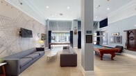 Located at 152 Temple in the heart of New Haven, CT, this single-floor community lounge offers a well-appointed gathering space designed for both relaxation and entertainment. The room is characterized by soaring ceilings adorned with crisp crown molding, recessed lighting, and light wood-style flooring that runs throughout, creating a bright and inviting atmosphere.

A defining architectural feature is the expansive floor-to-ceiling glazed storefront windows along one wall, dressed with navy blue drapes, which flood the interior with natural light and offer direct views of Temple Street. The lounge is divided into two distinct functional areas. The seating zone is anchored by a large New Haven city map wall mural, providing a locally inspired focal point, alongside comfortable gray sofas arranged around a marble-top coffee table, a wall-mounted flat-screen television, and accent lighting.

The adjacent game area centers on an ornate billiards table with richly carved legs and a slate-blue felt surface, complemented by a dedicated cue rack and framed artwork. A bar-height counter with stools runs along the window wall, ideal for casual gathering. Structural columns define the space without interrupting its open flow, while decorative touches — including a mounted deer head and vintage-style accessories — lend the lounge a distinctive, clubhouse-inspired character.