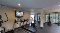 This fitness center is designed to provide a practical and comfortable exercise experience. The space is anchored by a row of TRUE-brand treadmills positioned prominently at the front, complemented by an elliptical trainer and additional cardio equipment toward the rear. A flat-screen television is wall-mounted at an ideal viewing height, allowing users to stay entertained during workouts. Recessed lighting throughout the ceiling provides consistent, even illumination across the space. The flooring features dark, durable material well-suited for a high-traffic fitness environment. Natural light enters through large windows at the far end of the room, creating an open and inviting atmosphere. Glass-paneled doors or partitions are visible toward the back, adding a sense of depth and separation while maintaining visual connectivity within the space. The neutral-toned walls contribute to a clean, focused aesthetic. The overall layout is thoughtfully organized, with equipment arranged to allow comfortable movement between machines, making this fitness center a functional and well-appointed facility for daily exercise.