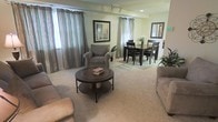 This single-floor residence presents an open-concept layout that seamlessly connects the living and dining areas, creating a spacious and functional environment. The living area is anchored by plush, neutral-toned wall-to-wall carpeting that extends throughout the space, contributing to a warm and inviting atmosphere. A round dark wood coffee table with a lower shelf serves as a central focal point, complemented by comfortable upholstered seating including a cushioned armchair and sofa.

Striped drapery panels in muted tones of teal and grey frame the windows, allowing natural light to filter softly into the room while adding a layered, decorative element to the walls. Track lighting along the ceiling provides additional ambient illumination. Framed wall art and a rectangular mirror add visual interest to the neutral-painted walls.

The dining area, positioned adjacent to the living space, features a dark wood rectangular dining table with matching chairs, accommodating everyday dining needs. A floral arrangement and additional greenery bring subtle natural accents to the space. The overall design maintains a cohesive, neutral palette that creates a calm and functional living environment.