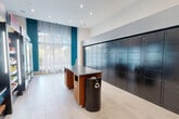 Located at 3400 Columbia Pike in Arlington, Virginia, this well-appointed mail and package room serves as a practical and modern amenity space. The room is finished with light-toned large-format tile flooring and features crisp white ceilings with recessed lighting that keeps the area bright and welcoming. A bold teal accent wall adds a contemporary touch, complemented by sheer floor-to-ceiling curtains that diffuse natural light from an exterior-facing window, offering a view of surrounding greenery.

Dominating one wall is an expansive bank of dark, matte-finish package lockers arranged in a grid pattern, providing secure, individual parcel storage across multiple sizes. A centrally positioned wood and dark-surface work counter with built-in storage offers a convenient surface for sorting mail and packages, accompanied by a recycling bin for added functionality.

The space also incorporates a compact convenience retail area, featuring fully stocked refrigerated display cases offering a wide variety of beverages, as well as shelving units carrying snacks and everyday essentials. A self-checkout kiosk allows for quick, independent purchases. An adjacent doorway with warm wood trim leads to a neatly organized open shelving unit, hinting at additional service or reception functions nearby. The overall design balances utility with a polished, modern aesthetic.