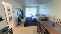 This single-floor residence features an open-concept layout that connects the dining and living areas, creating a cohesive and functional living space. The living room is anchored by a large window dressed with vertical blinds that allow an abundance of natural light to fill the room, lending the space a bright and airy feel. Light-colored walls complement the neutral carpeted flooring, while a deep navy area rug defines the seating zone and adds a bold contrast to the overall palette.

The living area is furnished with a generously sized grey sofa accented with coordinating cushions, a glass-topped coffee table, and a decorative accent chair with a patterned seat cushion. A floor lamp and wall-mounted mirror add layered lighting and decorative depth to the room. An entertainment unit with a flat-screen television occupies one corner, offering a practical media setup.

Adjacent to the living area, the dining space accommodates a wooden table and chairs, providing a dedicated area for meals. The textured ceiling adds architectural character throughout. Overall, the space presents a comfortable, well-lit, and cohesive interior suited for everyday living.