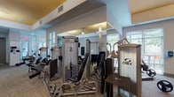 This fitness center offers a comprehensive workout environment designed to accommodate a wide range of training styles. The space is organized into distinct zones, each tailored to specific exercise needs. The cardio area is positioned along a wall of large windows that invite abundant natural light and outdoor views, housing multiple treadmills, ellipticals, and stationary bikes. Ceiling fans and warm gold-painted ceiling accents give this area a bright, energetic atmosphere.

The strength training zone is well-stocked with professional-grade Cybex weight machines, including cable systems, leg press, and various resistance stations. Floor-to-ceiling mirrors line one section of the space, enhancing the sense of openness while allowing users to monitor their form. A dedicated functional training area features TRX suspension trainers mounted to a wall-mounted rig, resistance bands, a BOSU ball, stability ball, and a balance board, providing versatile options for bodyweight and corrective exercise.

The flooring transitions between patterned carpet tile in the main exercise areas and dark rubberized flooring in the functional zone. A wall-mounted flat-screen television is positioned for visibility across the space. The center is accessed through a glass-panel entry door, and posted fitness center policies keep users informed of guidelines.