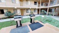 This inviting outdoor courtyard serves as the communal heart of a multi-story residential complex, offering a thoughtfully designed space for relaxation and outdoor living. At the center of the courtyard lies a sparkling swimming pool, surrounded by a fenced perimeter and framed by mature trees, tropical palms, and well-maintained ornamental shrubs that lend the area a lush, resort-like ambiance. The landscaping features layered planting beds with a variety of greenery, creating a natural buffer that enhances privacy and visual appeal throughout the space. A dedicated BBQ area is equipped with multiple stainless steel gas grills stationed on rubberized mats, providing a convenient and practical setup for outdoor cooking and entertaining. Adjacent to the grilling station, a shaded outdoor dining area features a glass-top table, cushioned chairs, and a large umbrella, ideal for al fresco meals in a comfortable, sun-protected setting. The surrounding building facades, finished in warm earth tones, feature private balconies on upper floors that overlook the courtyard below. Exterior stairwells provide access between levels, and ground-floor units open directly onto the shared outdoor space. The overall atmosphere is relaxed and community-oriented, with natural light and greenery contributing to a pleasant and welcoming outdoor environment.