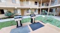 This inviting outdoor courtyard serves as the communal heart of a multi-story residential complex, offering a thoughtfully designed space for relaxation and outdoor living. At the center of the courtyard lies a sparkling swimming pool, surrounded by a fenced perimeter and framed by mature trees, tropical palms, and well-maintained ornamental shrubs that lend the area a lush, resort-like ambiance. The landscaping features layered planting beds with a variety of greenery, creating a natural buffer that enhances privacy and visual appeal throughout the space. A dedicated BBQ area is equipped with multiple stainless steel gas grills stationed on rubberized mats, providing a convenient and practical setup for outdoor cooking and entertaining. Adjacent to the grilling station, a shaded outdoor dining area features a glass-top table, cushioned chairs, and a large umbrella, ideal for al fresco meals in a comfortable, sun-protected setting. The surrounding building facades, finished in warm earth tones, feature private balconies on upper floors that overlook the courtyard below. Exterior stairwells provide access between levels, and ground-floor units open directly onto the shared outdoor space. The overall atmosphere is relaxed and community-oriented, with natural light and greenery contributing to a pleasant and welcoming outdoor environment.