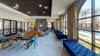 This single-floor community clubhouse is a thoughtfully designed social space that blends contemporary style with warm, inviting character. The main lounge is a standout area, featuring soaring ceilings with rich wood paneling, a series of dramatic arched windows dressed in golden drapery, and an abundance of natural light that frames views of the surrounding courtyard and community buildings. Seating throughout includes a tufted leather sofa, plush navy chaise lounges, and accent chairs arranged across patterned area rugs, creating multiple relaxed gathering zones anchored by a wall-mounted display screen.

Adjacent to the lounge is a spacious social kitchen and bar area, where a long white quartz island is lined with vibrant yellow wire barstools beneath a row of elegant glass pendant lights. The back wall features bold patterned tile work, open shelving, a mounted television, and stainless steel appliances, making it ideal for casual entertaining. A separate dining nook offers a high-top communal table with director-style chairs, pendant lighting, and Southwestern-inspired artwork set against a textured feature wall. Decorative accents throughout — including sculptural objects, woven curtains, and lush greenery — give the space a curated, resort-like ambiance.