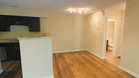This single-floor space offers a clean, functional layout designed for comfortable everyday living. The interior is anchored by warm-toned hardwood flooring that runs throughout, lending a sense of warmth and continuity to the space. Neutral, cream-colored walls create a bright and versatile backdrop, making the rooms feel open and inviting.

The living area benefits from a track lighting fixture mounted on the textured ceiling, providing focused illumination across the space. A half-wall partition separates the living area from what appears to be a kitchen or bar area, where a wall-mounted television and countertop surface are visible, adding both a functional and entertainment-oriented element to the layout.

The open-concept flow between the living and kitchen zones encourages a connected, sociable atmosphere. Crisp white baseboards frame the flooring and walls, adding a polished finishing detail throughout. A hallway visible in the background suggests additional rooms beyond the main living space, hinting at a thoughtful floor plan that balances openness with defined areas. Overall, the space presents a tidy, well-maintained interior with a straightforward aesthetic suited to a range of furnishing styles.