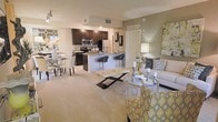 This single-floor residence is designed around an open-concept layout that connects the kitchen, dining, and living areas into one cohesive, flowing space. The kitchen is outfitted with dark espresso cabinetry, stainless steel appliances, and a granite-topped peninsula with bar seating for two, making it equally suited for casual meals and entertaining. Recessed lighting illuminates the kitchen and adjacent areas, contributing to a warm, inviting ambiance throughout.

The dining area, positioned just off the kitchen, accommodates a round glass-top table with upholstered seating, creating an intimate yet functional space for meals. The living area is generously proportioned, featuring plush neutral carpeting that extends across the main living zones. A glass-top coffee table and coordinating accent chairs in yellow and grey tones add a contemporary, layered aesthetic to the space.

Accent walls in a warm taupe tone provide subtle contrast against the lighter wall finishes, while a floor lamp and decorative artwork contribute to the overall polished atmosphere. A hallway leading from the living area suggests additional private spaces beyond. The open flow and cohesive design make this a practical and stylish single-floor residence.