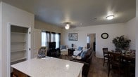 This single-story residence showcases an open-concept design that integrates the kitchen and living areas into one cohesive, functional space. The kitchen features a generous granite-look countertop that serves as both a workspace and a natural divider between the cooking and living zones, offering bar-style seating along its edge.

The living area is comfortably proportioned, accommodating a dark leather sofa and coordinating white armchairs accented with blue decorative pillows, creating a welcoming and cohesive aesthetic. A ceiling fan with a light fixture is centrally positioned overhead, providing both airflow and ambient lighting. Dark hardwood-style flooring runs throughout, adding warmth and continuity to the space.

Natural light enters through multiple windows dressed with dark curtain panels, while a flat-screen television is mounted on a media stand along one wall. Framed artwork and a decorative wall clock contribute subtle personality to the space. A hallway visible in the background suggests additional rooms beyond the main living area, reinforcing the home's practical, well-organized layout.