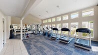 This single-story community clubhouse is designed to serve a variety of resident needs, combining fitness, leisure, and productivity in one cohesive space. The fitness center is the standout feature, characterized by soaring vaulted ceilings and an expansive bank of large windows that flood the room with natural light and offer views of the surrounding outdoor landscape. The gym is well-appointed with True-brand treadmills, elliptical machines, a multi-station cable machine, a dumbbell rack, and a medicine ball station, all set atop a blue area rug over tile flooring. Full-length mirrors along one wall enhance the sense of space. Adjacent to the fitness area, a casual lounge features a striking floor-to-ceiling brick fireplace with a wall-mounted television above, providing a warm and inviting atmosphere. A dining area nearby offers round tables with modern chairs beneath recessed lighting, with natural light entering through white-shuttered windows. The community kitchen includes warm wood cabinetry, stone-patterned countertops, stainless steel appliances, and tile flooring. A compact business center workstation with a computer, printer, and decorative mirror rounds out the amenities.