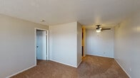 This single-story unit is laid out with a practical, straightforward floor plan that connects the main living area, kitchen, and bedrooms in a cohesive arrangement. Throughout the carpeted areas, warm-toned, medium-pile carpet runs consistently, providing a unified look across the living room, bedrooms, and hallways. The textured ceilings are present throughout the unit, adding a classic residential character.

The living room is a generously sized space featuring a ceiling fan with a light fixture and a large window fitted with horizontal blinds, allowing natural light to filter in. A front entry door opens directly into this area. An interior half-wall divides the living space, offering a sense of separation while maintaining an open feel.

The kitchen is designed in a galley configuration, featuring dark wood upper and lower cabinetry, light-colored countertops, and a yellow tile floor. It is equipped with a refrigerator, stovetop, and dishwasher, with a door at the far end providing additional access.

The bedrooms are well-proportioned, each featuring windows with blinds for natural light and privacy. One bedroom includes a wall-mounted HVAC unit beneath its window. Closet access and a bathroom connection are visible in the primary bedroom, which also benefits from two doorways for convenient circulation throughout the unit.