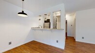 This well-appointed single-floor residence makes excellent use of its intimate footprint, offering a cohesive layout that includes a living room, bedroom, kitchen, bathroom, two closets, and a hallway. Warm hardwood flooring flows throughout the main living areas, lending continuity and a welcoming character to the space.

The living room is a bright, open area anchored by a large picture window that draws in natural light and a ceiling fan overhead, creating a comfortable and airy atmosphere. Adjacent to the living space, the kitchen features a semi-open pass-through design with a breakfast bar, pendant lighting, stainless steel appliances including a full-size refrigerator, dark cabinetry, and a striking mosaic tile backsplash that adds visual interest.

The bedroom is a well-proportioned retreat with its own ceiling fan, a window for natural light, and access to a closet. The bathroom is tastefully finished with a white vanity featuring a marble-style countertop, mosaic tile backsplash that mirrors the kitchen, large-format floor and wall tiles, and a full soaking tub with an overhead shower. Two closets provide practical storage solutions throughout the unit. Overall, the space presents a clean, modern aesthetic with thoughtful design details.