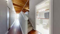 This loft-style space showcases a compelling blend of industrial character and modern comfort throughout its single-floor layout. A defining architectural feature is the exposed wood beam ceiling, which spans the main living areas and corridors, adding warmth and a raw, industrial aesthetic that contrasts beautifully with the neutral gray walls beneath.

Rich hardwood flooring runs throughout the primary living spaces, reflecting natural light and enhancing the sense of openness. A carpeted staircase with a clean white railing connects levels within the unit, positioned alongside a hallway that leads to the bathroom. Tall, multi-pane windows in one of the rooms flood the space with natural light and frame urban skyline views, further reinforcing the loft's city-dwelling character.

The galley-style kitchen is well-appointed with warm cherry wood cabinetry, dark granite countertops, and a full suite of black appliances including a range, refrigerator, dishwasher, and over-the-range microwave. The full bathroom features white tile flooring, a tub-shower combination with white tile surround, a white vanity with under-cabinet storage, a wood-framed mirror, and bright vanity lighting. Exposed ductwork in the living area adds to the space's industrial appeal.