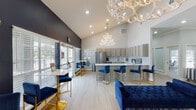 Located at 3802 Caravelle Pkwy in Corpus Christi, Texas, this single-story clubhouse space makes a striking first impression with its bold navy blue and crisp white color palette carried throughout. The entry reception area welcomes visitors with a sleek, modern white tiered front desk, light wood-look flooring, crown molding, and recessed lighting. A large abstract artwork anchors the accent wall, setting a sophisticated tone from the moment you walk in.

The open lounge area flows seamlessly from the reception, featuring soaring vaulted ceilings adorned with dramatic white antler chandeliers that serve as the defining design centerpiece of the space. Deep navy blue tufted velvet sofas and benches with gold-accented legs are arranged throughout, complemented by coordinating bar-height chairs and bistro tables positioned near large windows that invite natural light and views of the outdoor area.

The fully equipped kitchen is both functional and refined, offering grey shaker-style cabinetry, white quartz countertops with a peninsula bar, stainless steel appliances including a full-size refrigerator and microwave, and a decorative tile backsplash. The kitchen opens directly to the lounge, making it ideal for community gatherings and events. Glass doors provide convenient access to an outdoor pool area, enhancing the overall sense of openness and connectivity throughout the space.
