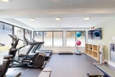 The Pulse Fitness Centre, located at 2650 41st Avenue East in Vancouver, BC, is a thoughtfully designed exercise facility offering a comprehensive range of equipment for a full-body workout experience. The space is bathed in natural light, courtesy of large, frosted windows that line the walls, creating a bright and energizing atmosphere while maintaining privacy. Recessed circular ceiling lights provide consistent, even illumination throughout.

The facility is divided into distinct workout zones. The cardio area features multiple treadmills and an elliptical trainer, positioned along the windowed wall to take advantage of natural light. A multi-functional cable machine and adjustable weight bench anchor the strength-training section, complemented by a full dumbbell rack with an expansive wall mirror that enhances the sense of space. Functional training accessories, including stability balls, a BOSU balance trainer, and step platforms, are neatly organized near a built-in shelving unit.

The flooring transitions between durable rubber sport tiles in the main exercise zone and a smooth concrete finish near the entry area, which also houses a water bottle filling station. Soft green accent walls near the entry add a calming touch. A wall-mounted flat-screen TV provides entertainment, making The Pulse a well-rounded, amenity-rich fitness centre.