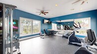 This dedicated fitness center offers a fully functional workout environment designed to accommodate a range of exercise needs. The space is defined by bold teal-painted walls contrasted against crisp white trim and ceilings, creating an energetic and motivating atmosphere. Durable speckled rubber flooring covers the entire area, providing a safe and resilient surface for all types of training activities.

The room is outfitted with a comprehensive selection of equipment, including a multi-functional cable machine, a treadmill, an elliptical, a stationary bike, and a weight bench paired with a tiered dumbbell rack. A full-length wall mirror with a wooden ledge runs along one side of the room, visually expanding the space while allowing users to monitor their form during workouts.

Natural light fills the room through multiple large windows positioned on multiple walls, offering views of the surrounding outdoor environment. A ceiling fan with integrated lighting aids in climate comfort and air circulation throughout the space. A wall-mounted flat-screen television provides entertainment during workouts. Recessed lighting supplements the natural light, ensuring the room remains bright at all times. The fitness center is accessed through a paneled door, making it a self-contained and purpose-built facility.