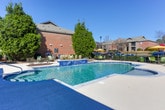 Located in Phenix City, Alabama, this outdoor community amenity area centers around a generously sized swimming pool with clear, blue-tinted water. The pool features clearly marked depth indicators, a built-in water feature along one edge, and a curved zero-entry spa section equipped with stainless steel handrails, making it accessible and inviting. Blue tile accents line portions of the pool's perimeter, adding a polished decorative detail.

The surrounding concrete sundeck is expansive, providing ample space for relaxation. Multiple green sling-style lounge chairs and tables are arranged throughout the deck, with vibrant yellow patio umbrellas offering shaded seating areas. The deck is enclosed by a wrought iron fence, providing both security and an unobstructed view of the surrounding brick residential buildings and mature landscaping, including a prominent evergreen tree that serves as a natural focal point.

Adjacent to the pool, a covered pavilion structure features a pitched roof and floor-to-ceiling glass panel walls with sliding door access, offering a sheltered outdoor gathering space. The area benefits from abundant natural light under open skies, creating a bright and welcoming atmosphere well-suited for outdoor recreation and leisure.