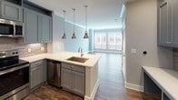 This 1-bedroom, 1-bathroom residence is laid out entirely on a single floor, offering a cohesive and efficient living environment with contemporary finishes throughout. The kitchen serves as a central highlight, featuring grey shaker-style cabinetry that extends to the ceiling, crisp white quartz countertops, a stainless steel undermount sink set within a peninsula island, and a full suite of stainless steel appliances including a refrigerator, range, and microwave. A tile backsplash adds texture and visual interest, while three brushed nickel pendant lights hang above the island, providing both task lighting and a stylish design accent. Rich hardwood-style flooring flows through the kitchen and into the open living area, where large windows fitted with horizontal blinds allow generous natural light to fill the space. The bedroom is carpeted for comfort and features a ceiling fan, a large window with outdoor views, and neutral grey walls that lend a calm, restful atmosphere. The bathroom is well-proportioned and refined, showcasing large-format tile flooring, grey vanity cabinetry with a white countertop, a wide mirror with vanity lighting, and a glass-enclosed walk-in shower with floor-to-ceiling tile surround. Soft, neutral tones and consistent modern finishes tie the entire residence together cohesively.