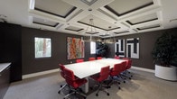 This professional conference room is designed to impress, combining elegant architectural details with bold, contemporary aesthetics. The standout feature of the space is its ornate coffered ceiling, framed in white molding against a dark surround, which adds depth and a sense of grandeur to the room. Suspended from the ceiling are two large, circular brushed-steel pendant light fixtures that provide focused, ambient illumination across the room.

The centerpiece of the space is a long, sleek white conference table surrounded by high-backed red leather executive chairs on polished chrome bases, creating a striking visual contrast against the deep charcoal-toned walls. The walls serve as a gallery for bold contemporary artworks, including a vibrant abstract splatter painting and a geometric black-and-white piece, adding creative energy to the professional environment.

Along one end of the room, dark wood credenza cabinetry provides functional storage, accompanied by a wall-mounted flat-screen display suitable for presentations. A glass-paneled door offers a refined entry point, with sidelights allowing a glimpse into adjacent spaces. Windows along the perimeter provide natural light and views of lush tropical foliage outside. Neutral carpet flooring ties the design together, balancing the room's bold color palette with understated elegance.