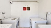 Located in Deerfield Beach, Florida, this shared utility room serves as a dedicated laundry facility for the building's residents. The space is compact yet efficiently organized, housing multiple commercial-grade washing machines and dryers arranged along the walls. The machines include LG Commercial Washer W11 Direct Drive units, reflecting a functional, no-frills setup designed for regular shared use.

The interior features light wood-style flooring that contrasts with the bright white walls, keeping the space feeling clean and open. An electrical panel is mounted on one wall, and exposed plumbing connections run along the back wall, supporting the multiple appliances. Posted laundry room rules signs are prominently displayed, providing clear usage guidelines for residents.

A full-width doorway opens directly to an outdoor courtyard, allowing natural light and fresh Florida air to flow into the room. The courtyard view reveals a well-maintained grassy area surrounded by the building's multi-unit residential structure, offering a pleasant backdrop to an otherwise utilitarian space.

The room's single-floor layout maximizes the available footprint to accommodate the laundry equipment while maintaining ease of movement. Overall, this facility provides a straightforward, accessible, and practical laundry solution for the building's residents.